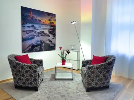 Therapieraum mit zwei gemusterten Sesseln, kleinem Tisch mit Orchidee und großem Meeresbild an der Wand